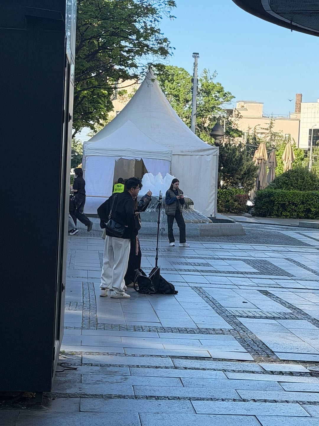 Two people with a tripod set up in a busy Belgrade plaza near a fountain and mall entrance.