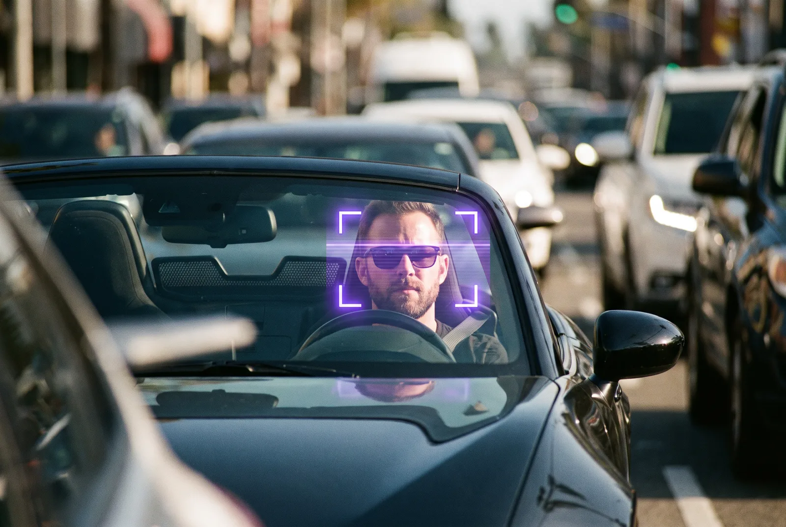 A daytime view zoomed onto the driver of an open-top convertible stopped in traffic, the driver's face framed by a violet facial-recognition detection box.